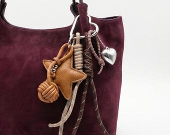 May include: A burgundy suede tote bag with a silver heart-shaped clasp and a collection of charms. Charms include a brown leather star, a knotted leather ball, and a silver heart pendant. The bag has a soft, textured appearance.