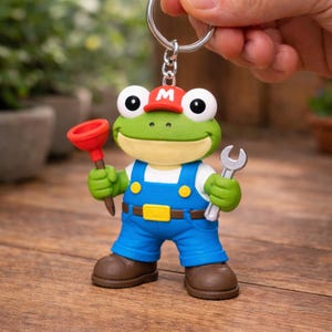 Puede incluir: Un llavero novedoso con un personaje de rana de dibujos animados que lleva un mono azul, una gorra roja con una "M" blanca y botas marrones. La rana sostiene un desatascador rojo y una llave inglesa plateada. El llavero está unido a un anillo plateado.