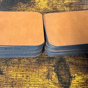 May include: Two stacks of rectangular coasters. Each coaster has a light brown top and dark grey sides. The coasters are stacked on a wooden surface with a warm, brown tone.