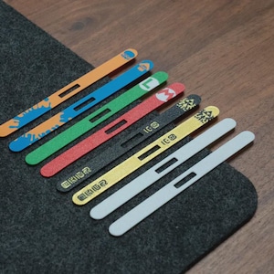 May include: A collection of colorful, flat, rectangular game controller accessories. The accessories are orange, blue, green, red, black, yellow, and white. Some have character designs and text. They are arranged on a dark gray surface.