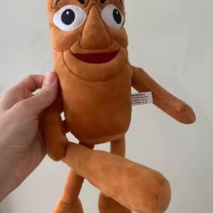 May include: A plush toy in the shape of a cartoon character with a brown body, arms, and legs. The toy has a large nose, wide eyes, and a smiling mouth. The toy is being held by a hand.