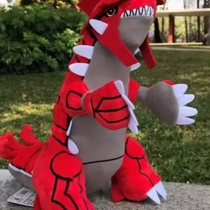 May include: A red and grey plush toy creature with white accents. The toy features a large head with a red crest, white teeth, and a grey body. It has large red arms and legs with white claws. The toy is positioned on a stone surface.