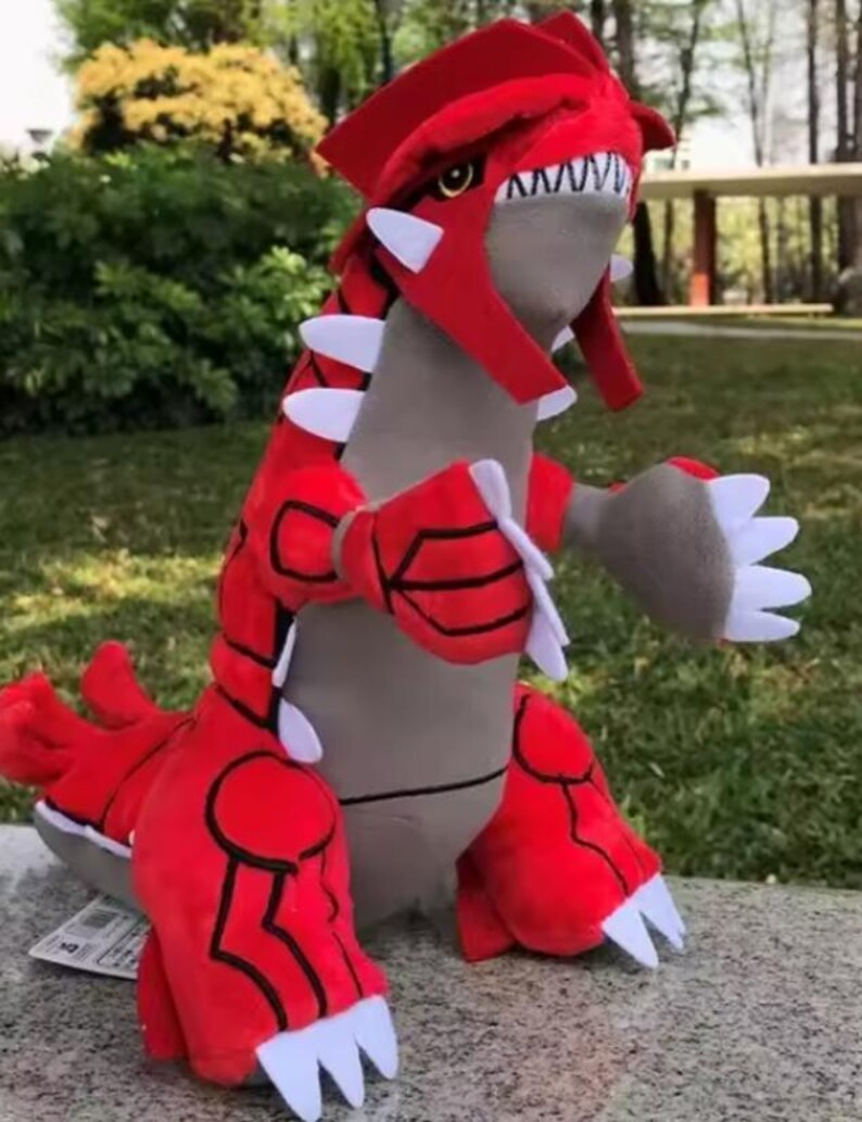 May include: A red and grey plush toy creature with white accents. The toy features a large head with a red crest, white teeth, and a grey body. It has large red arms and legs with white claws. The toy is positioned on a stone surface.
