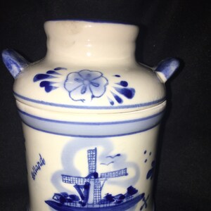 May include: A white and blue ceramic container with a lid and two handles. The container is decorated with a windmill scene and the word "Holland". The lid features a blue floral design.
