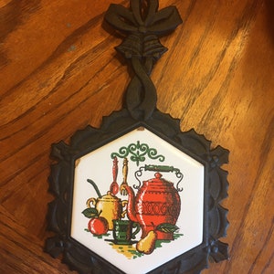 Könnte beinhalten: Schwarzer gusseiserner Topfuntersetzer mit sechseckiger Fliese mit einer Küchenszene mit roter Teekanne, gelber Zuckerdose und Obst. Der Untersetzer hat ein dekoratives Bogen- und Schriftrollen-Design.