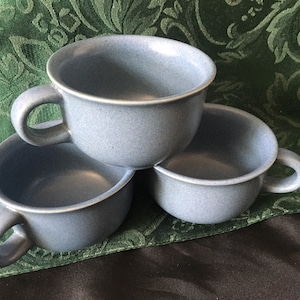 以下が含まれることがあります： 取っ手付きの青い斑点模様のセラミックカップが3つ積み重ねられています。カップは丸みを帯びた形状で、シンプルなデザインです。カップはくすんだ青色で、斑点模様のテクスチャがあります。カップはおそらく飲み物を提供するためのものです。