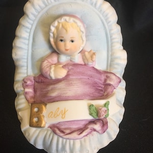 May include: A ceramic figurine of a baby in a pale blue cradle. The baby wears a pink bonnet and outfit, with a purple blanket. A banner with the word "Baby" is across the front.