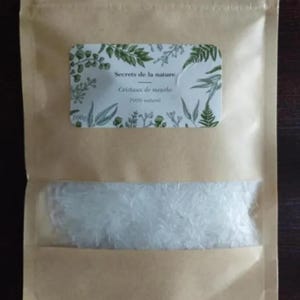 Puede incluir: Una bolsa de papel marrón con una ventana transparente que muestra cristales blancos. Una etiqueta rectangular con ilustraciones botánicas verdes y el texto "Secrets de la nature Cristaux de menthe 100% naturel" está adherida.