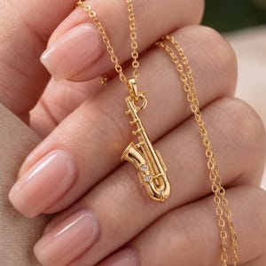 Peut inclure: Un collier doré avec un pendentif en forme de saxophone. Le pendentif est orné de deux petites pierres rondes et transparentes incrustées dans le corps de l'instrument. Le bijou est suspendu à une fine chaîne à maillons dorée assortie.