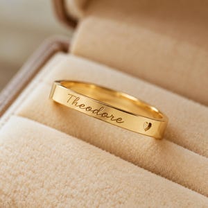 Könnte beinhalten: Ein polierter goldfarbener Ring mit dem eingravierten Namen Theodore in Schreibschrift, ergänzt durch ein kleines eingraviertes Herzsymbol, das sich rechts neben dem Namen auf der Außenseite des Metallrings befindet.