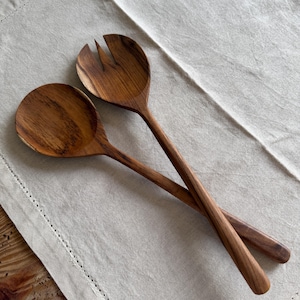 May include: Two wooden salad servers, a spoon and a fork, are crossed on a light beige linen surface. The spoon has a round bowl, and the fork has three tines. The wood has a warm, natural grain.