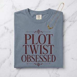 Puede incluir: Una camiseta de manga corta color azul pizarra con cuello redondo y etiqueta Comfort Colors. El frente presenta texto en color granate oscuro que dice PLOT TWIST OBSESSED con adornos decorativos. Un collar de cadena dorada descansa sobre la tela.