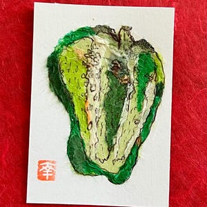 May include: A rectangular white card featuring a textured paper collage of a halved green bell pepper. The artwork uses various shades of green and white paper layers with black ink outlines. A small red square stamp with the Japanese character 傘 is visible.