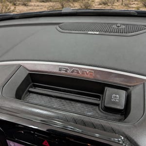 May include: A black vehicle dashboard featuring a RAM logo on a metallic trim piece, a recessed storage tray with a textured base, and a 12V DC power outlet cover. The dashboard is positioned below a windscreen showing a desert landscape.
