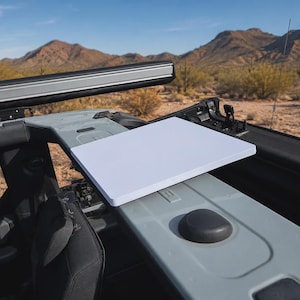May include: A rectangular white flat surface tray mounted onto the interior frame of a vehicle. The tray is positioned horizontally over a gray plastic console component featuring a circular black knob, with a desert landscape visible outside.
