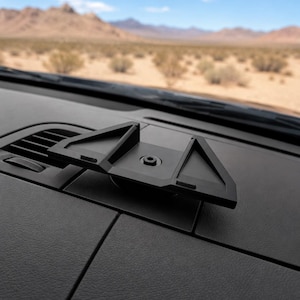 May include: A black plastic vehicle dashboard mount featuring a triangular frame design with a central circular attachment point. The mount is secured to the flat surface of a car dashboard, positioned near the air vent and windshield area.