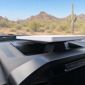 May include: A white, rectangular, flat-panel antenna mounted on a black pedestal base, positioned securely on the dashboard of a vehicle. The device features a clean, minimalist design with a smooth top surface and a tapered support structure.