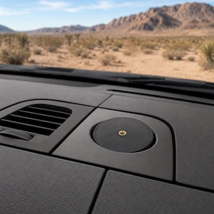 May include: A circular black mounting base with a centered brass-colored threaded insert, integrated into a dark gray textured vehicle dashboard surface next to a rectangular air vent, designed for securing electronic device accessories.