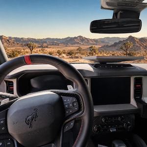 May include: The interior of a vehicle featuring a black steering wheel with a red centre stripe and a horse emblem. The dashboard includes a central touchscreen, climate controls, and the word BRONCO in red lettering on the passenger side.