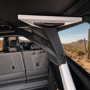 May include: A white rectangular shelf mounted to the interior roll bar of a vehicle. The shelf is supported by a black angled bracket attached to the frame. The vehicle interior features black textured panels, seats, and a view of a desert landscape.