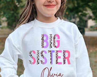 May include: A white long-sleeve sweatshirt featuring the text BIG SISTER in a mix of pink, purple, and leopard print patterns. Below the main text, the name Olivia is printed in a dark red cursive script font on the front chest.
