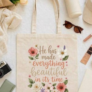 May include: A beige canvas tote bag featuring a floral design and the text "He has made everything beautiful in its time" followed by "Ecclesiastes 3:11". The graphic includes various colourful flowers and green leaves surrounding the printed lettering.