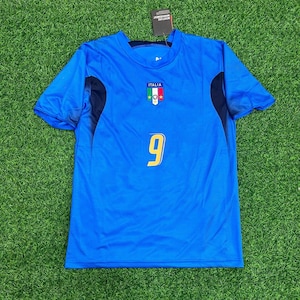 以下が含まれることがあります： イタリア国旗のエンブレムと黄色の9番が付いた青いサッカーユニフォーム。肩には濃い青色のアクセントがあり、上部にはタグが付いています。