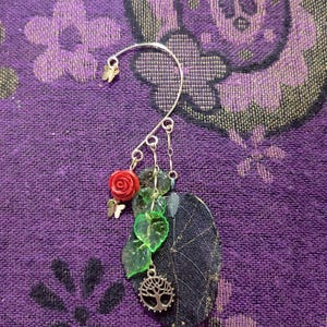 May include: A silver ear cuff featuring a red rose, green leaves, and a dark leaf pendant. The cuff includes a small butterfly charm and a tree of life pendant, creating a nature-inspired design. The cuff is set against a purple patterned background.