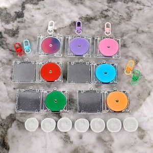 May include: A selection of clear acrylic CD-shaped keyrings with colourful inserts in shades of pink, purple, blue, red, green, and orange. Each keyring has a silver clasp. Additional small keyrings and clear plastic discs are also present.