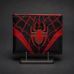 May include: Black wallet with a red and black spiderweb design and a red spider emblem. The wallet is displayed on a silver stand against a dark background. The wallet has a bi-fold design.