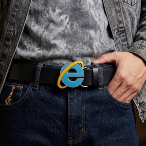 Peut inclure: Une ceinture en cuir noir avec une boucle représentant le logo bleu, jaune et blanc d'Internet Explorer. La ceinture est portée avec un jean bleu et une veste en jean grise. La boucle est au centre de l'image.