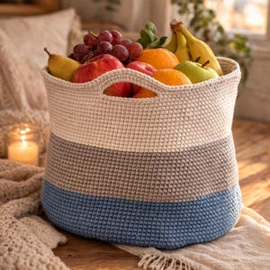 Peut inclure: Un panier en crochet fait à la main, crème, gris et bleu, contient une variété de fruits frais, notamment des pommes, des oranges, des raisins, des poires et des bananes. Le panier a deux poignées et est posé sur une surface en bois.