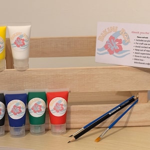 Puede incluir: Una caja de madera con seis tubos de pintura acrílica amarilla, blanca, negra, azul, verde y roja. Cada tubo tiene una etiqueta floral. También hay dos pinceles y una tarjeta con texto.