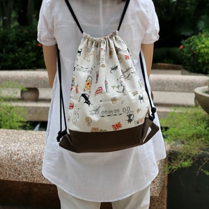 May include: A white drawstring backpack with a brown bottom. The backpack has a black and white print of cats, flags, and other designs. The backpack is being worn by a person.