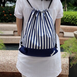 Pode incluir: Uma mochila de cordão de cor branca e azul marinho com a parte inferior de cor azul marinho. A mochila está sendo usada por uma pessoa.