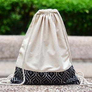 May include: A white drawstring bag with a black and white geometric pattern around the bottom. The bag is made of cotton and has a simple design.