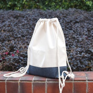 Puede incluir: Una bolsa de cordón blanco con un fondo de mezclilla azul. La bolsa tiene un cordón blanco y está hecha de una tela suave y natural.