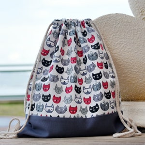 Può includere: Una borsa a cordoncino grigia con sfondo bianco e un motivo di gatti neri e rossi. La borsa ha un fondo grigio e cordoncini bianchi.