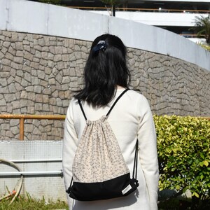 Könnte beinhalten: Eine Person trägt einen weißen Pullover mit einem schwarzen und weißen floralen Rucksack mit Kordelzug.