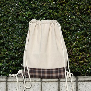 Puede incluir: Una bolsa de cordón blanco con un ribete de cuadros marrones y negros. La bolsa está hecha de un tejido suave y natural y tiene un diseño simple y clásico.