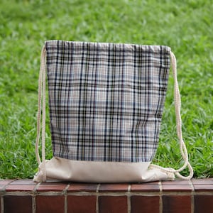 Puede incluir: Una bolsa de cordón hecha de tela blanca con un patrón de cuadros negros, blancos y azules. La bolsa tiene un fondo de algodón blanco y cordones blancos.