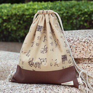 Puede incluir: Una bolsa de cordón beige con detalles marrones. La bolsa está hecha de tela y tiene un diseño de caracteres chinos negros. La bolsa tiene un fondo marrón y un cordón blanco. La bolsa tiene una etiqueta que dice "silverbreeze".
