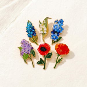 Könnte beinhalten: Eine Sammlung von sechs floralen Emaille-Anstecknadeln. Die Anstecknadeln zeigen detaillierte Designs verschiedener Blumen, darunter blaue Hyazinthen, Maiglöckchen, Vergissmeinnicht, lila Flieder und rote Mohnblumen. Jede Anstecknadel hat eine goldfarbene Umrandung und grüne Blätter.