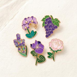 Op de afbeelding: Vijf emaille pins met bloemen- en fruitontwerpen. De pins bevatten een paarse orchidee, een tros druiven, lavendel, een viooltje en een roze pioenroos. Elke pin heeft goudkleurige contouren en groene bladeren.