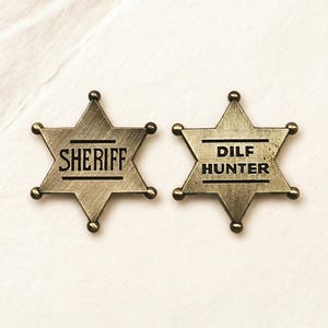 Könnte beinhalten: Zwei sternförmige Abzeichen im Antik-Stil mit Bronze-Finish. Auf einem Abzeichen steht "SHERIFF" und auf dem anderen "DILF HUNTER" in schwarzen Großbuchstaben. Jeder Sternpunkt hat eine kleine, runde, bronzefarbene Verzierung.