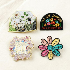 Peut inclure: Quatre épingles émaillées avec différents motifs. L'une représente une serre avec des plantes, une autre dit "I've got sunshine on a cloudy day" avec des fleurs, une troisième dit "Shameless Smut Lover", et la dernière est une fleur avec des affirmations positives.