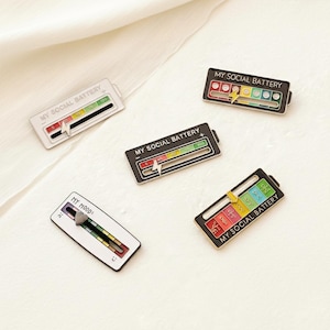 May include: Enamel pins with the text "MY SOCIAL BATTERY" and "MY MOOD". The pins feature a battery-like design with a lightning bolt and a sliding scale, displaying levels from low to high. The pins are in various colors.