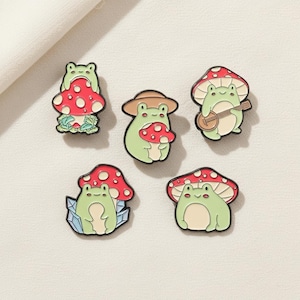 May include: Set of five enamel pins featuring whimsical frog designs. Each pin showcases a green frog with a mushroom, some with hats or musical instruments. The mushrooms are red with white spots, and the pins have a black outline.