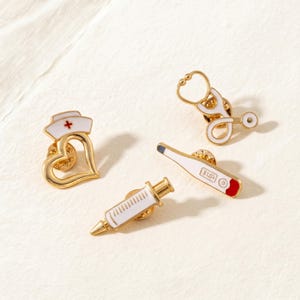 May include: Four gold-colored medical-themed pins. The pins include a nurse's hat on a heart, a stethoscope, a thermometer showing 31.0 degrees, and a syringe. The pins have white and red enamel details.