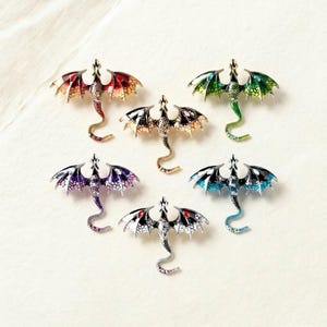 Könnte beinhalten: Eine Sammlung von sechs Drachenbroschen in verschiedenen Farben. Jede Brosche zeigt ein detailliertes Drachendesign mit ausgebreiteten Flügeln und einem eingerollten Schwanz. Die Drachen sind in Rot-, Grün-, Lila-, Schwarz- und Blautönen mit metallischen Akzenten gehalten.